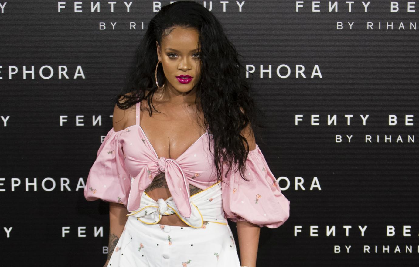 Rihanna wears a shoulder and cleavage baring pink top tied with a center knot paired with a white print skirt, big hoop earrings, and long wavy hair at the 2017 Fenty Beauty Launch event.