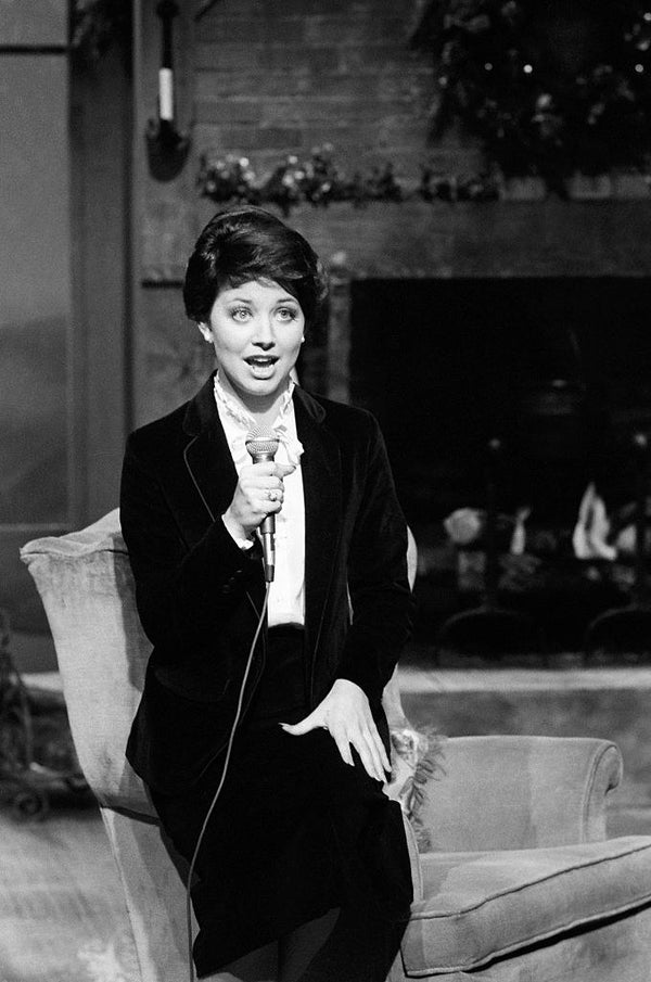 Powell holding a mic and sitting on an armchair