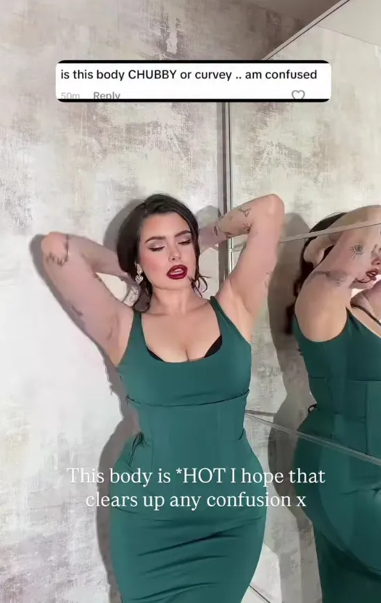 A woman in a green dress and dark red lipstick poses with her arms above her head, next to a mirror that reflects her.